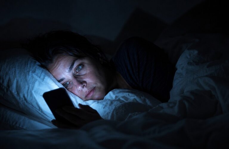 Person awake in bed at 3am illuminated by smartphone screen glow, representing digital insomnia and compulsive news consumption