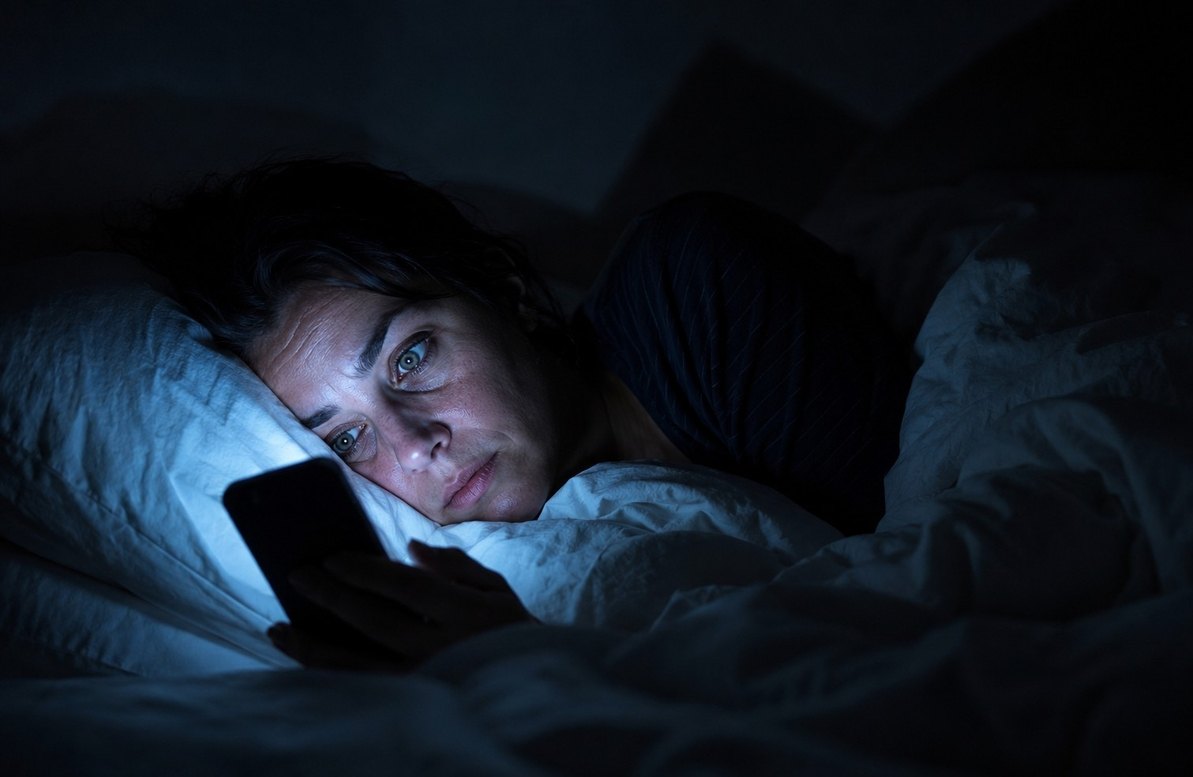 Person awake in bed at 3am illuminated by smartphone screen glow, representing digital insomnia and compulsive news consumption